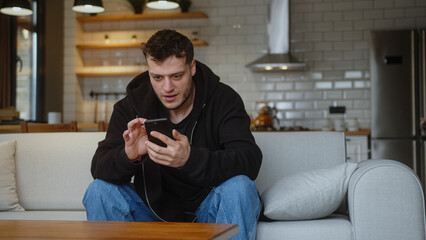  Young man sitting on sofa reacts to what sees, surfing internet on mobile phone, checking email, reading media news, scrolling social medias, using mobile applications on smartphone at home