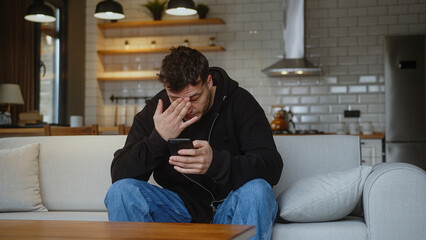 Young man sitting on sofa having headache while using smartphone. Tired young man get having pain...