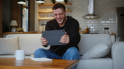 Overjoyed man playing a video game on a tablet computer while sitting on the sofa at home. Excited...