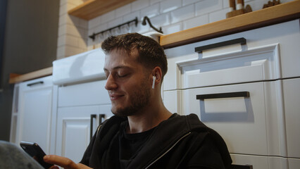 Carefree young man sitting on the kitchen floor listening to music on his smartphone with wireless...