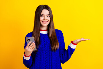 Photo portrait of pretty teen girl device palm hold empty space wear trendy knitwear blue outfit isolated on yellow color background