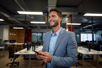 Happy young caucasian man in businesswear smiling while using mobile phone in office