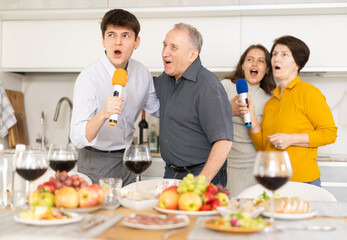 Cheerful lively multi-generational family, elderly parents and young adult children, singing into microphones, enjoying karaoke session at home dinner party with food and wine
