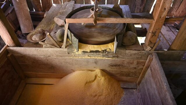 A millstone is spinning. A stone grinds grain in a mill. Production of corn flour. Corn kernels fall into the mill. Traditional watermill on the river.