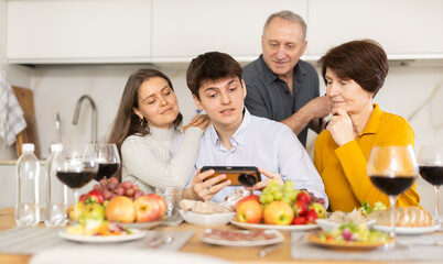 Positive young couple sharing memories with elderly parents sitting at cozy kitchen, browsing photos on smartphone together during festive family meal