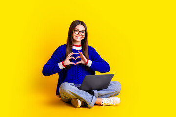 Full length photo of pretty teen girl hold netbook heart crossed legs wear trendy knitwear blue outfit isolated on yellow color background