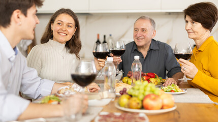 Obraz na płótnie Canvas Positive daughter with boy-friend or husband and parents friendly conversation at dinner table with wine and light snacks in cozy kitchen
