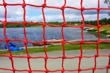 Close-up of a red knotted fishing net with a serene lake and colorful boats in the distant...