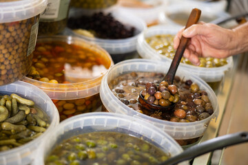 In self-service store, mens hands hold ladle and place pickled black olives with stone in...