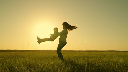 Mother and child have fun in meadow, mother rotates her daughter, baby flies, laughs, rejoices. Family walk in nature. Happy family, mom, little daughter play together in park on green grass in sun.