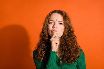 Photo of uncertain doubtful lady dressed green shirt hand chin emtpy space isolated orange color background