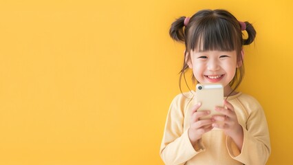 Cute asian child using smart phone and smiling, standing over pastel yellow wall, copyspace