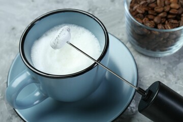 Mini mixer (milk frother), whipped milk in cup and coffee beans on grey textured table, closeup