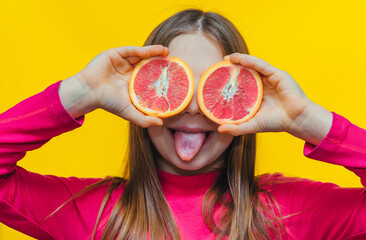 Little cute funny girl uses grapefruit instead of eyes, shows her tongue and has fun in the studio on a yellow background. Concept of citrus diet, fruits and vitamins.