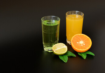 Two glasses with different citrus juices on a black background, next to half a lime and an orange with leaves.