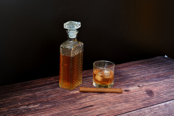 A glass of whiskey with ice, a crystal decanter and a Cuban cigar on a dark wooden table.