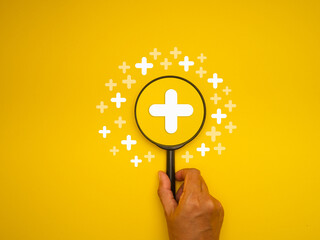 Hand holding a magnifying glass against a yellow background with white plus symbols.