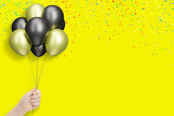 Female hand holding bunch of shiny golden and black balloons on yellow background with confetti....