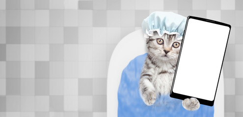 Cute kitten wearing shower cap takes the bath at home and shows big smartphone with white blank...