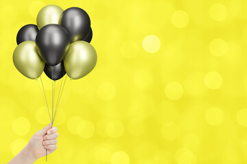 Female hand holds bunch of golden and black balloons on blurred yellow background. Empty space for text