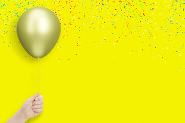 Female hand holds golden balloon on yellow background with confetti. Empty space for text