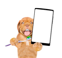 Mastiff puppy with funny big teeth holds the toothbrush with toothpaste and holds big smartphone with white blank screen in it paw. isolated on white background