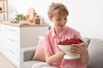 Beautiful happy young woman with colander full of ripe cherries sitting on sofa in kitchen - obrazy, fototapety, plakaty