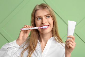 Young woman brushing her teeth and holding tube of toothpaste on green background