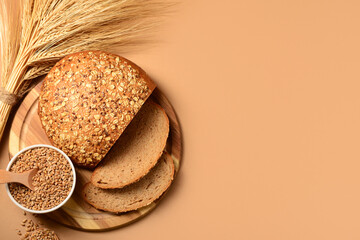 Wooden board with sliced loaf of bread, wheat spikelets and grains on beige background