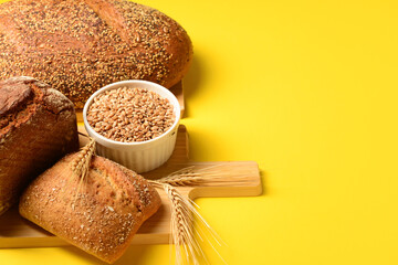 Wooden board with loaves of fresh bread, wheat spikelets and grains on yellow background