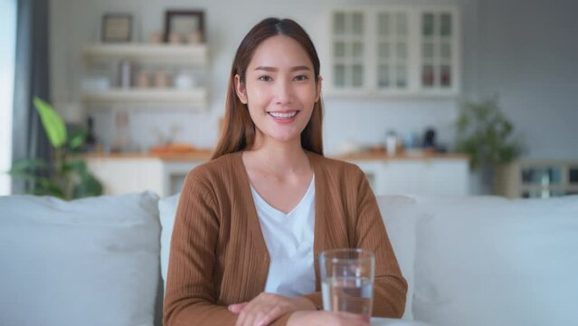 Young Asian woman smiling looking at camera sitting on sofa happy expression, hand holding water glass
