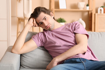 Weary young man sitting on sofa at home - obrazy, fototapety, plakaty