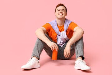 Young man with can of soda sitting on pink background