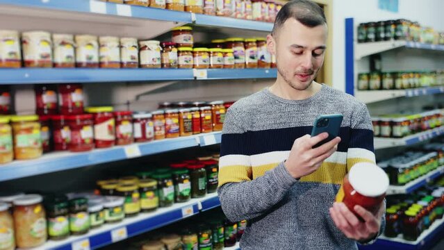 Man in store of Russian goods photographs jar canned lecho - stewed salad made from bell pepper, tomato sauce. Buyer scan QR code compares item in store and in mobile application 