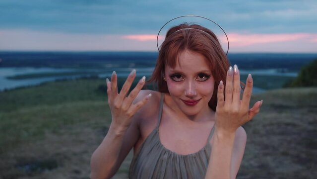 A red-haired girl with a halo on her head moves her hands gently in the air and looks directly into the camera. Medium shot. Slow motion. 4k