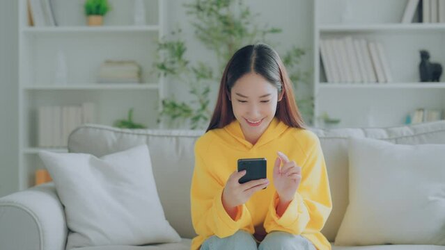 Happy young Asian woman relax sitting on couch using cell phone, smiling lady laughing holding smartphone, looking at cellphone enjoying doing online ecommerce shopping in mobile apps