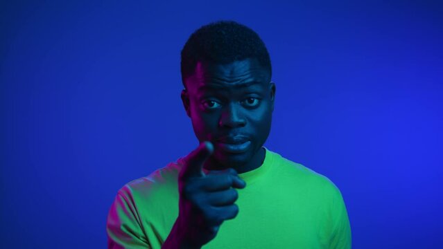 Portrait of Black Man Looking at Camera with Pointing Finger Gesture Inside Neon Room. Modern Stress Lifestyle of One African American Close-Up. Anger in Argue of Fashionable 20s Person with Dark Skin