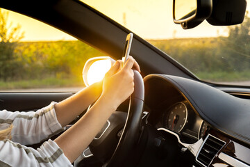 Young woman using mobile phone while driving car on highway road during sunset. Womandriver has...