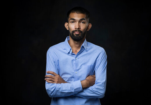 Portrait of young Asian man with beard in a blue shirt with his arms crossed and looking at camera in black studio background.