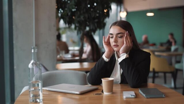 tired, beautiful woman sits in a stylish cafe in business attire, closing her laptop and finishing work with a headache, massaging her temples to relieve migraine pain