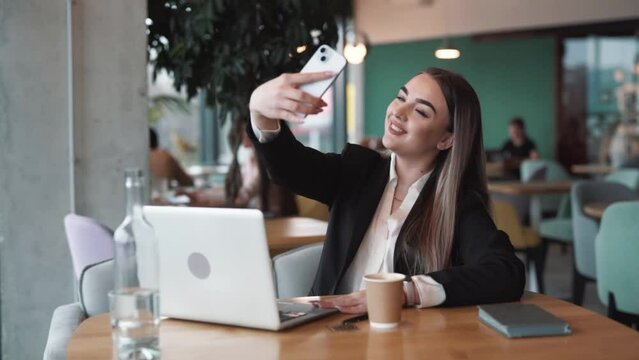 Beautiful woman sitting in a cafe in business attire takes a selfie with a smile on her face on a smartphone