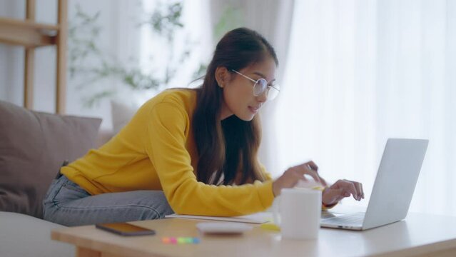 Young Asian female wearing glasses using laptop, working at home in living room, coffee mug on table. Cozy office workplace, remote work