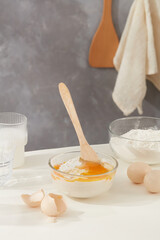 Eggs and flour are whipped with a wooden spoon placed on white kitchen counter against marble background, some ingredients surrounded. Cooking concept with front view