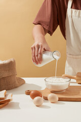 Fototapeta na wymiar Female hand is holding a milk glass bottle pours milk into a glass bowl next to ingredients for homemade bakery on light brown background. Milk and egg has many benefits for human healths