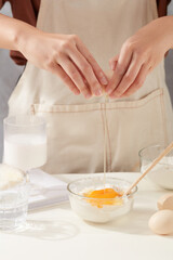 Baking ingredients concepts with female hands are cracking an egg in a bowl of flour to prepare homemade pastry on minimals white kitchen table next to ingredients. Front view photo
