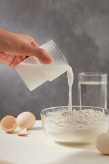 Marble background against culinary baking process in the kitchen with a mug hold by female man pours into a glass flour bowl beside eggs and glass of water on white kitchen table. 
