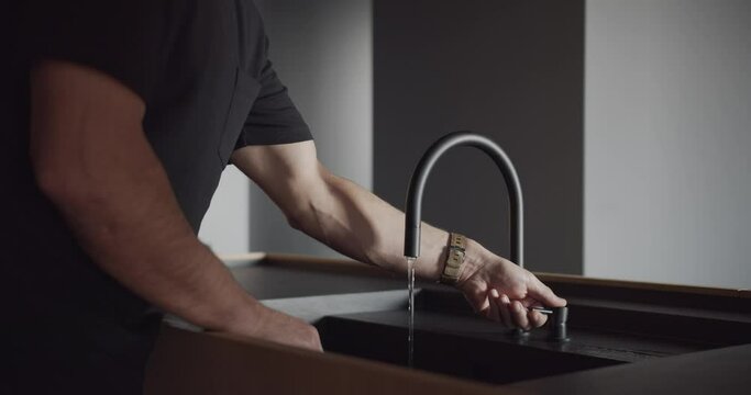 A man is washing his hands in the kitchen sink, using his thumb to turn on the faucet. The flooring is made of hardwood, a composite material
