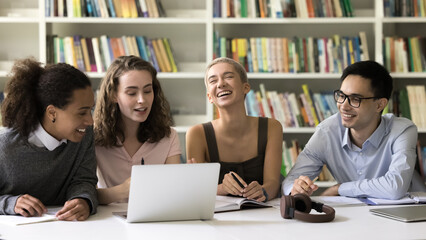Laughing interracial students gathered in university library, sit at desk with laptop, watch...