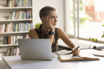 Thoughtful student girl sitting at desk with textbook and laptop, looks out window, deep in...
