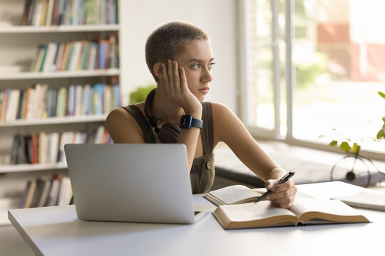 Thoughtful student girl sitting at desk with textbook and laptop, looks out window, deep in thoughts, thinking on mathematical problem, feels tired or unmotivated, disinterested in studying process